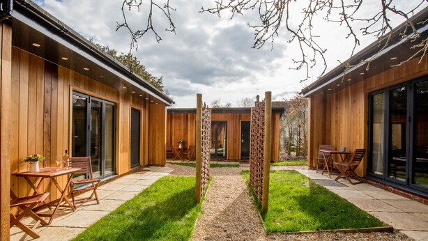 A modern building with wooden exterior walls, large sliding glass doors, and a gravel pathway leading to a central brick pillar. - The Thomas Lord (Petersfield)