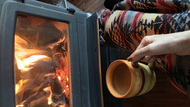 Une personne tenant une tasse près d’un poêle à bois.