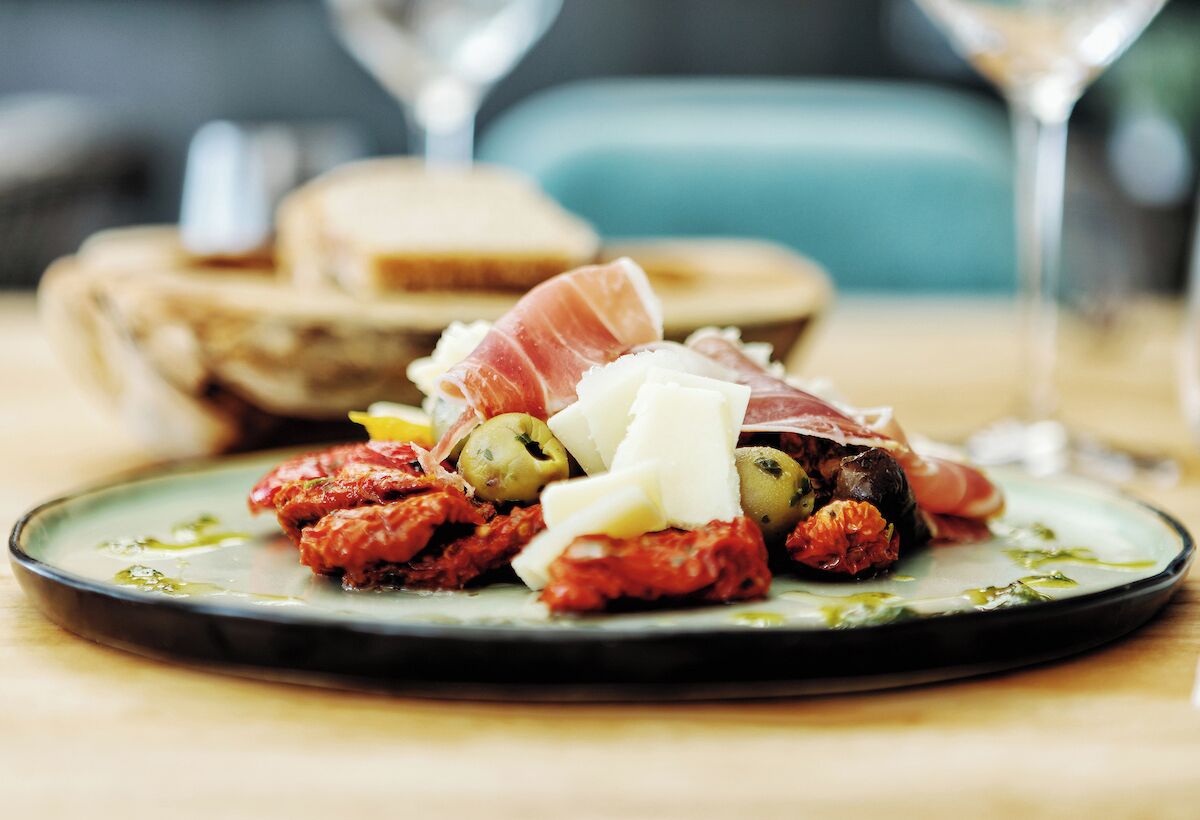 Un plato con una selección de embutidos, aceitunas y queso, sobre una mesa con una copa de vino al fondo.