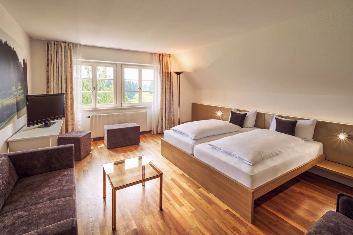 A modern hotel room with a large bed, a desk with a flat-screen TV, a small table, and a couch.