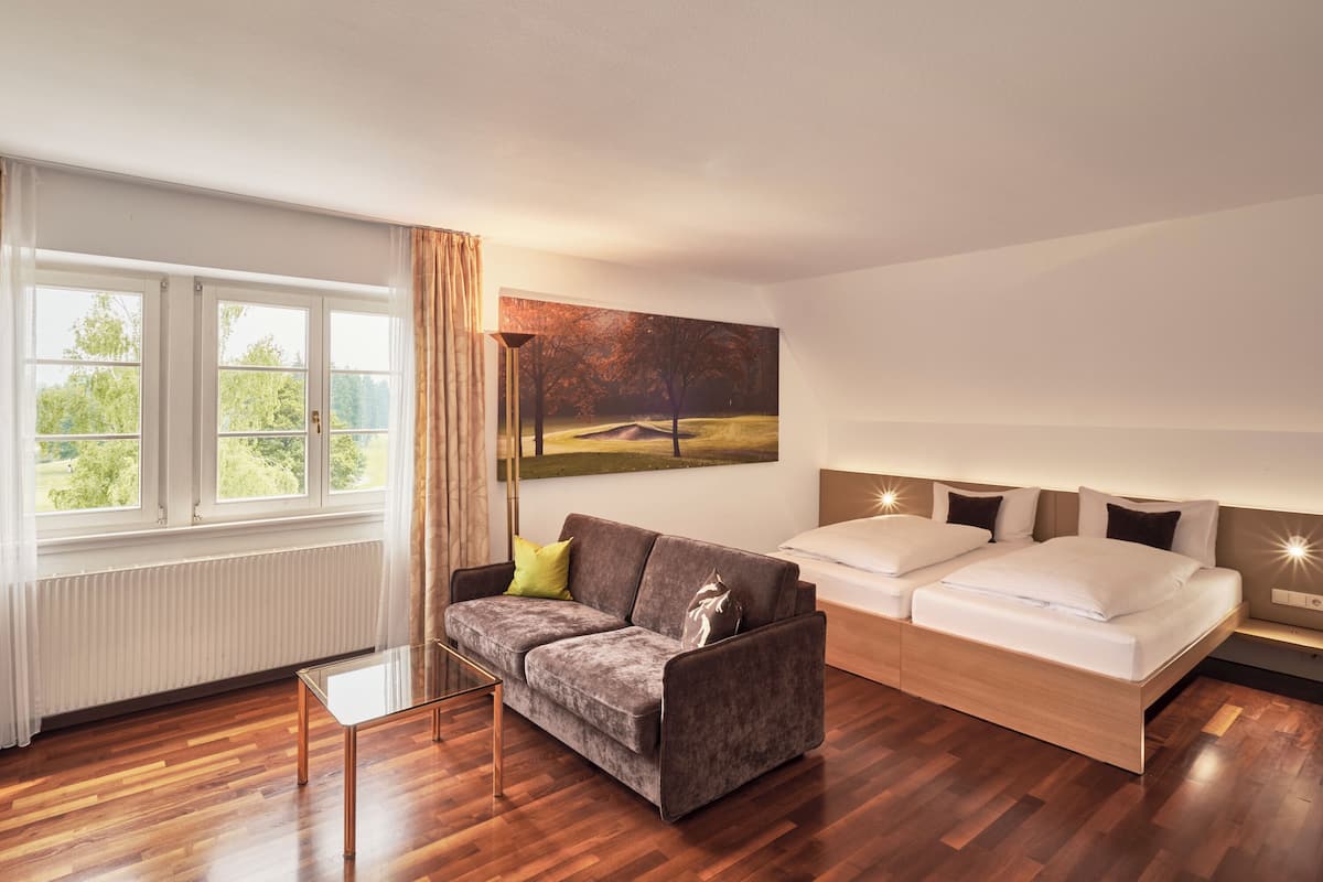 A modern hotel room with a wooden floor, a grey sofa, a bed with white bedding, and a large window with curtains.