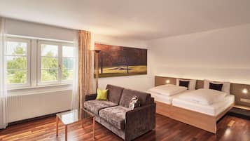 Una habitación de hotel moderna con suelo de madera, un sofá gris, una cama con ropa de cama blanca y un ventanal con cortinas.