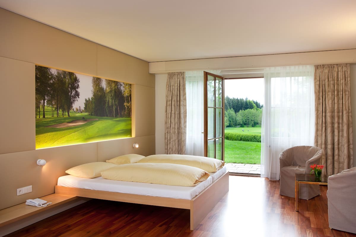 A modern bedroom with a bed, a chair, a small table with a vase, and a large window with curtains.