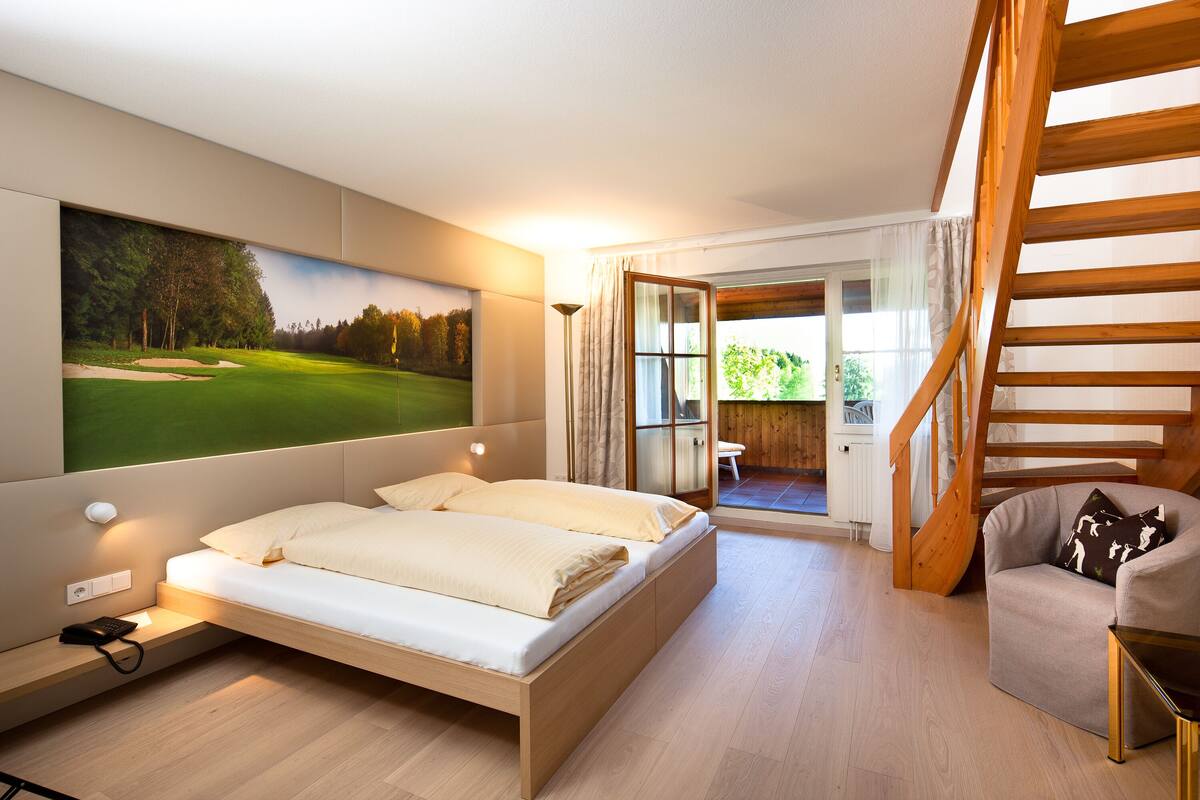 A bedroom with a bed, a desk, a chair, a staircase, and a view of a golf course.