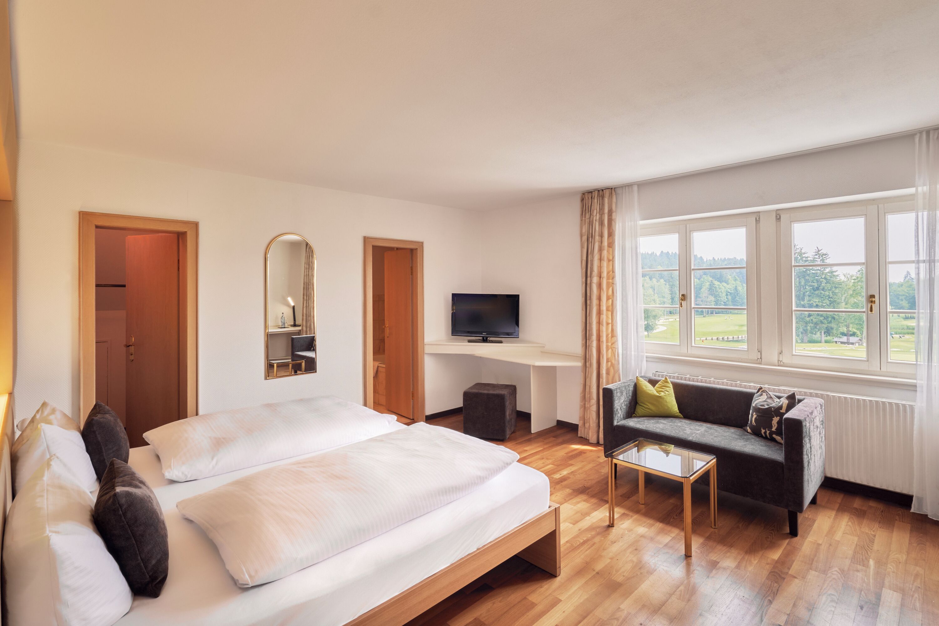 Una camera d'albergo con un letto, un divano, una televisione e una finestra vista verde.