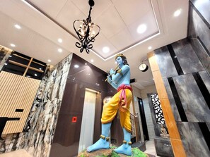 A statue of a blue-skinned figure with a flute, standing on a rock base in a modern lobby with marble walls and a chandelier. - Hotel Krishnas  (Amritsar)