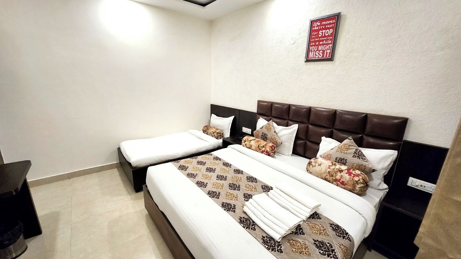 A hotel room with two single beds and a larger bed. There is a motivational quote on the wall and a small table with a lamp.