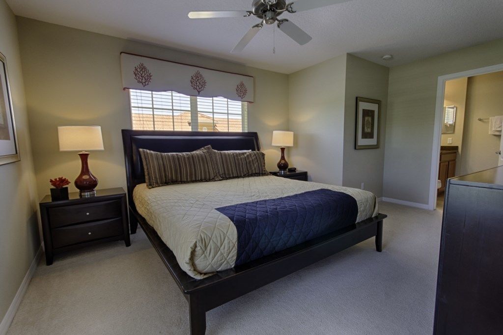 A bedroom with a bed, two bedside tables, a ceiling fan, a window with blinds, and a framed picture on the wall.