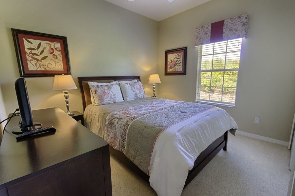 A bedroom with a bed, two bedside lamps, a desk with a computer, and a window with blinds.