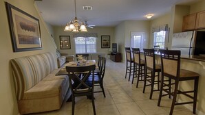 A dining area with a long table and chairs, a sofa, and a kitchen area with a refrigerator and cabinets.