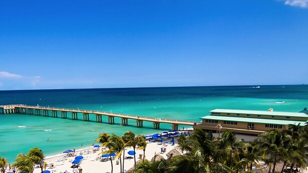 A beach with a pier extending into the ocean, a building with a green roof, and palm trees. - Modern 2BR/2BA Ocean View Beach Condo STR-09902 (Sunny Isles Beach)