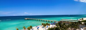 A beach with a pier extending into the ocean, a building with a green roof, and palm trees. - Modern 2BR/2BA Ocean View Beach Condo STR-09902 (Sunny Isles Beach)