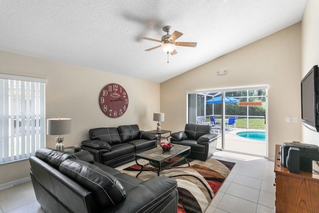 A spacious living room with a large clock, a ceiling fan, and a view of a swimming pool.