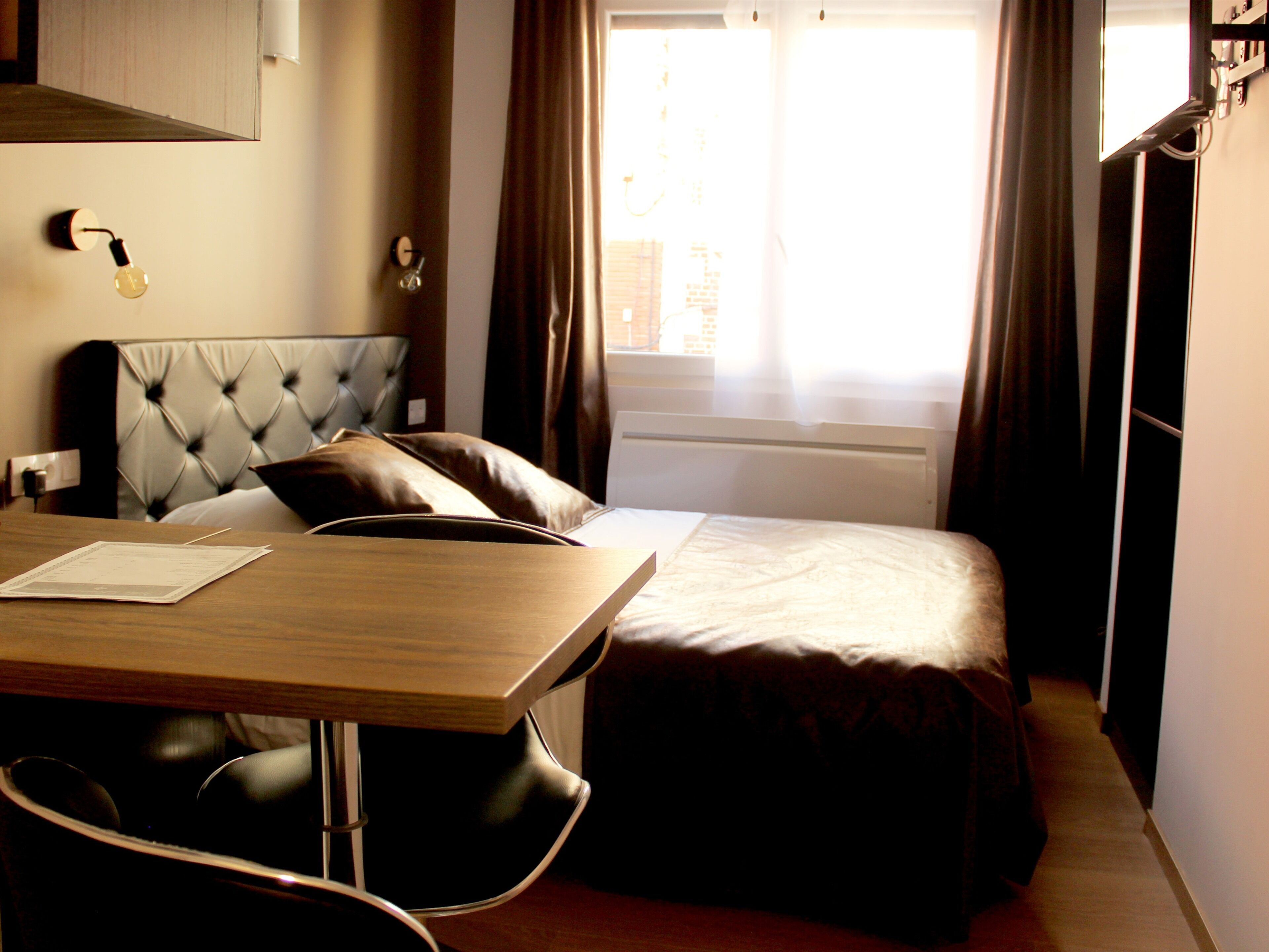 A hotel room with a bed, a bedside table, a chair, a window with curtains, and a wall-mounted light fixture.