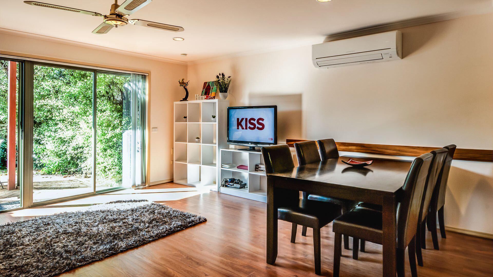 Un espace repas moderne avec une table et des chaises en bois, un téléviseur diffusant des images du groupe « KISS », et une étagère blanche.