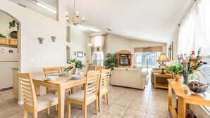 Ein geräumiger Wohnbereich mit Esstisch, Stühlen und einer Couch. Es gibt eine Küchenzeile mit Schränken und einem Kühlschrank. Der Raum verfügt über große Fenster und eine Kristallleuchter.