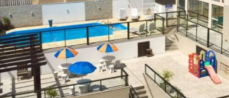 A rooftop pool area with a clear view of the pool, lounge chairs, umbrellas, and a children's play area.