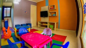 Una sala de juegos infantil con una mesa rosa, paredes azules y naranja, y un televisor en la pared.