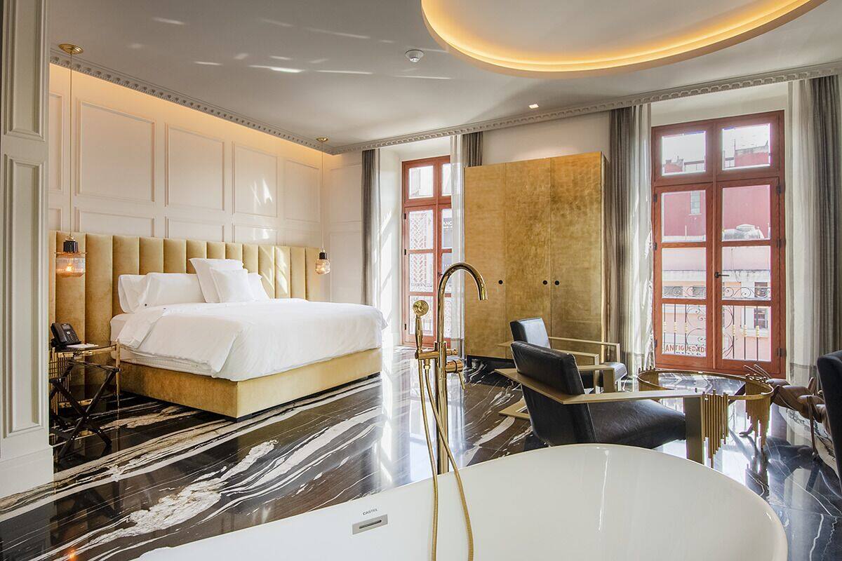 A hotel room with a large bed, a sitting area with a table and chairs, a bathtub, and a view of the city through large windows.