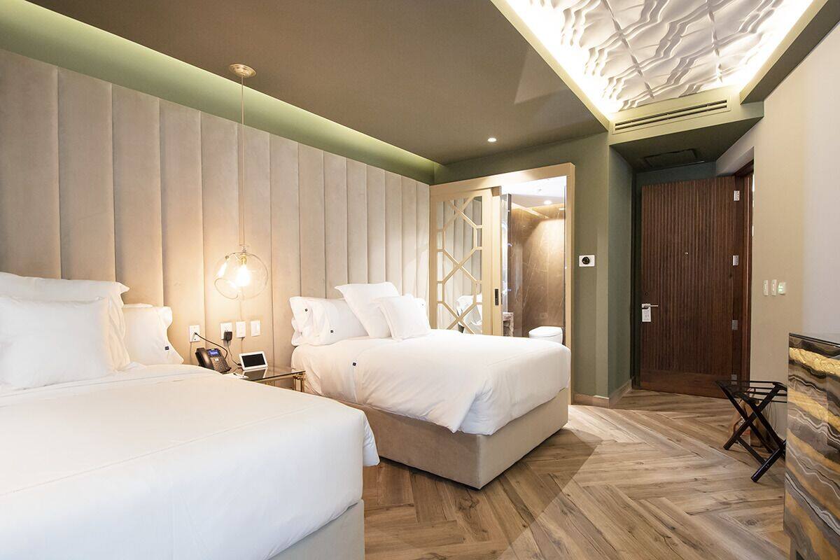 A hotel room with two beds, a wooden floor, a green accent wall, and a ceiling light fixture.