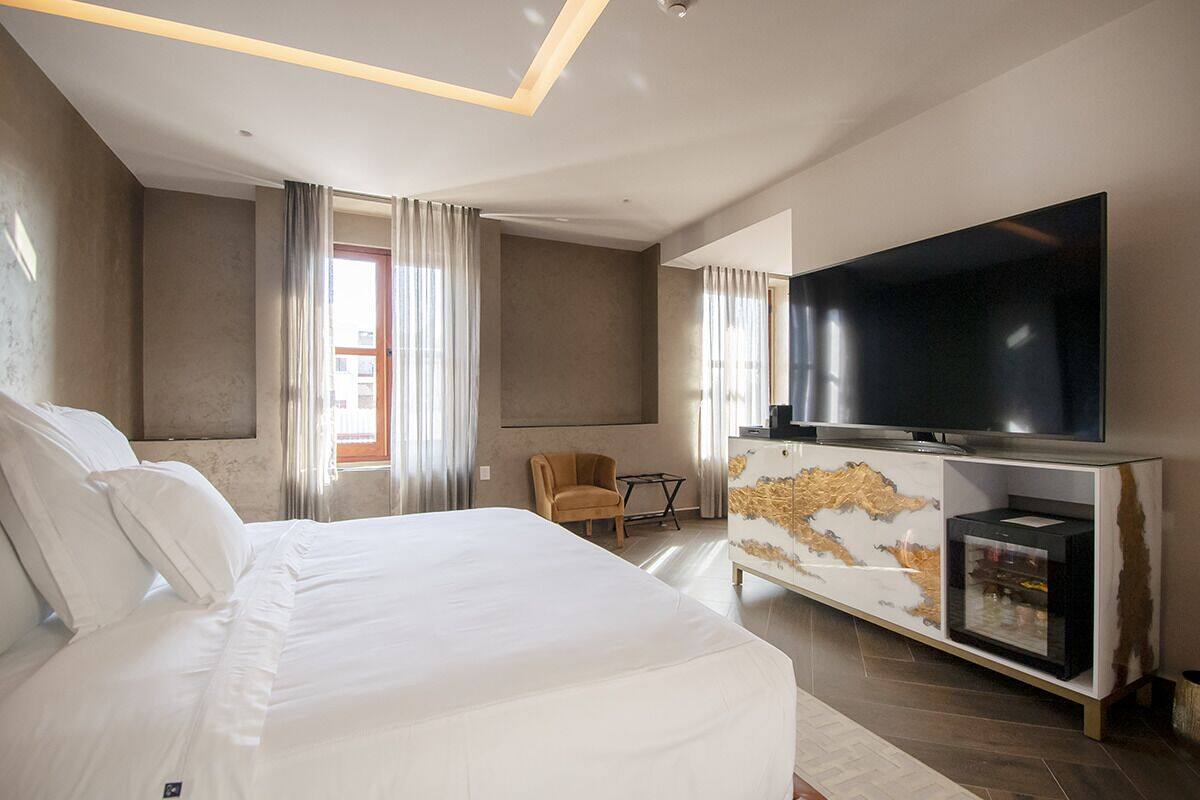 A modern hotel room with a large bed, a flat-screen TV, a minibar, and a seating area with a chair and table.
