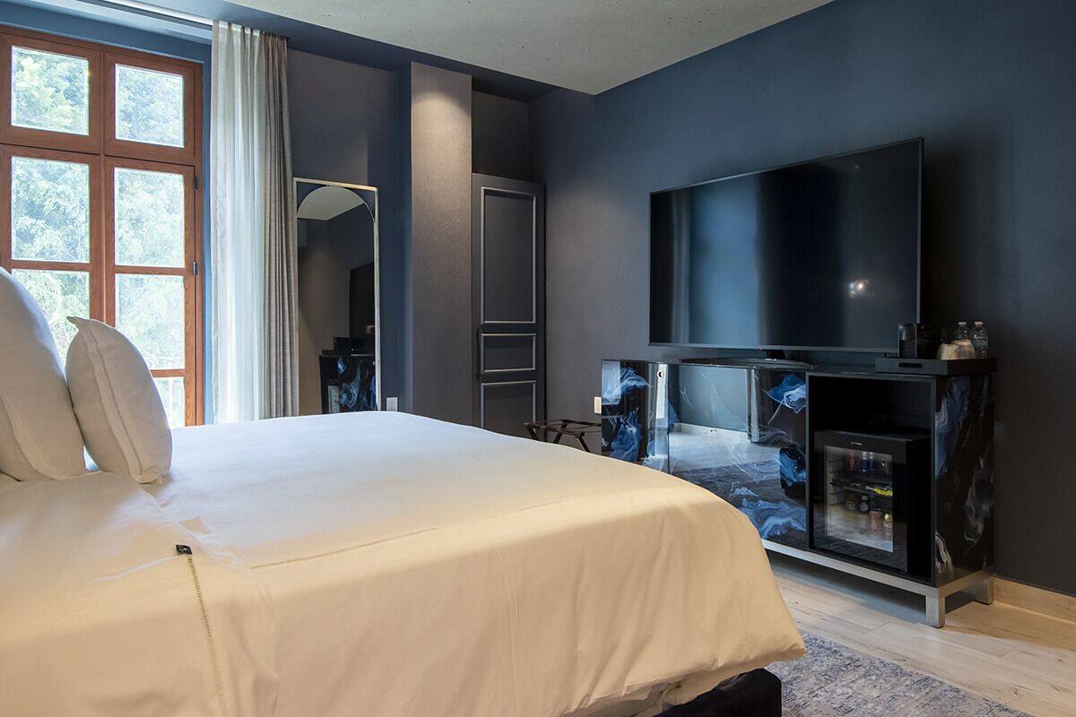 A modern hotel room with a large bed, a flat-screen TV, a marble-patterned console table, and a large window with curtains.