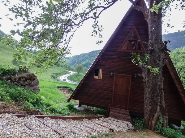 Una cabaña de madera con techo triangular, una ventana pequeña y una puerta. Está situada junto a un árbol y un sendero de grava.