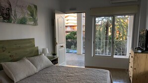 Une chambre à coucher comprenant un lit, une table de chevet, une commode et une fenêtre donnant sur un immeuble et de la verdure.