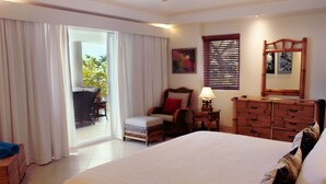 A hotel room with a large bed, a chair, a dresser, and a window with blinds. - Palm Beach Condo 202  - A Vacation Rental by Bougainvillea Barbados (Hastings)