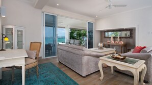 A spacious living room with a sofa, a coffee table, and a dining area with a table and chairs. There is a sliding glass door leading to a balcony with a view of the ocean.