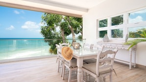 A dining area with a glass table and white chairs, overlooking a beach and ocean.