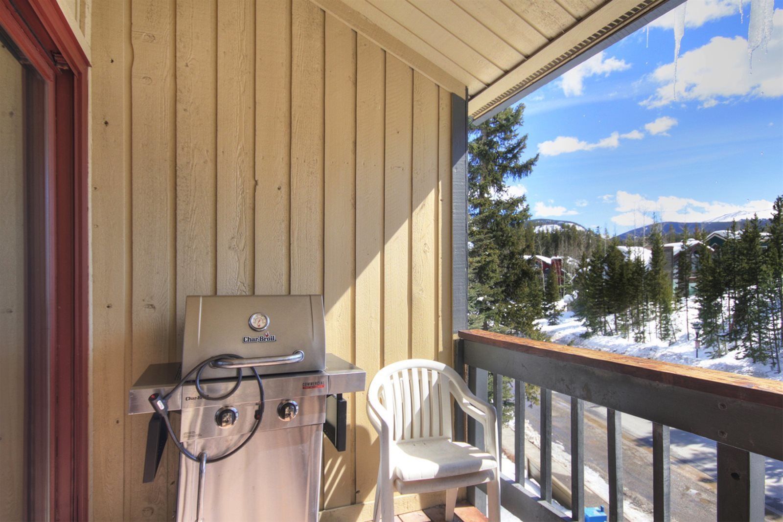 A balcony with a Char-Broil grill, a white plastic chair, and a view of a snowy landscape with trees and buildings.
