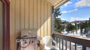 A balcony with a Char-Broil grill, a white plastic chair, and a view of a snowy landscape with trees and buildings.