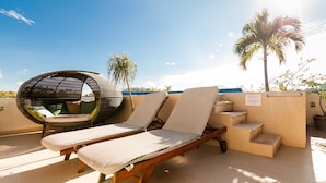 A rooftop lounge area with a wicker chair and two lounge chairs, a palm tree, and a view of the ocean. - Kingsize 1bdr apt, common rooftop w/ lounge area, pool, BBQ... (units 202) (Playa Del Carmen)