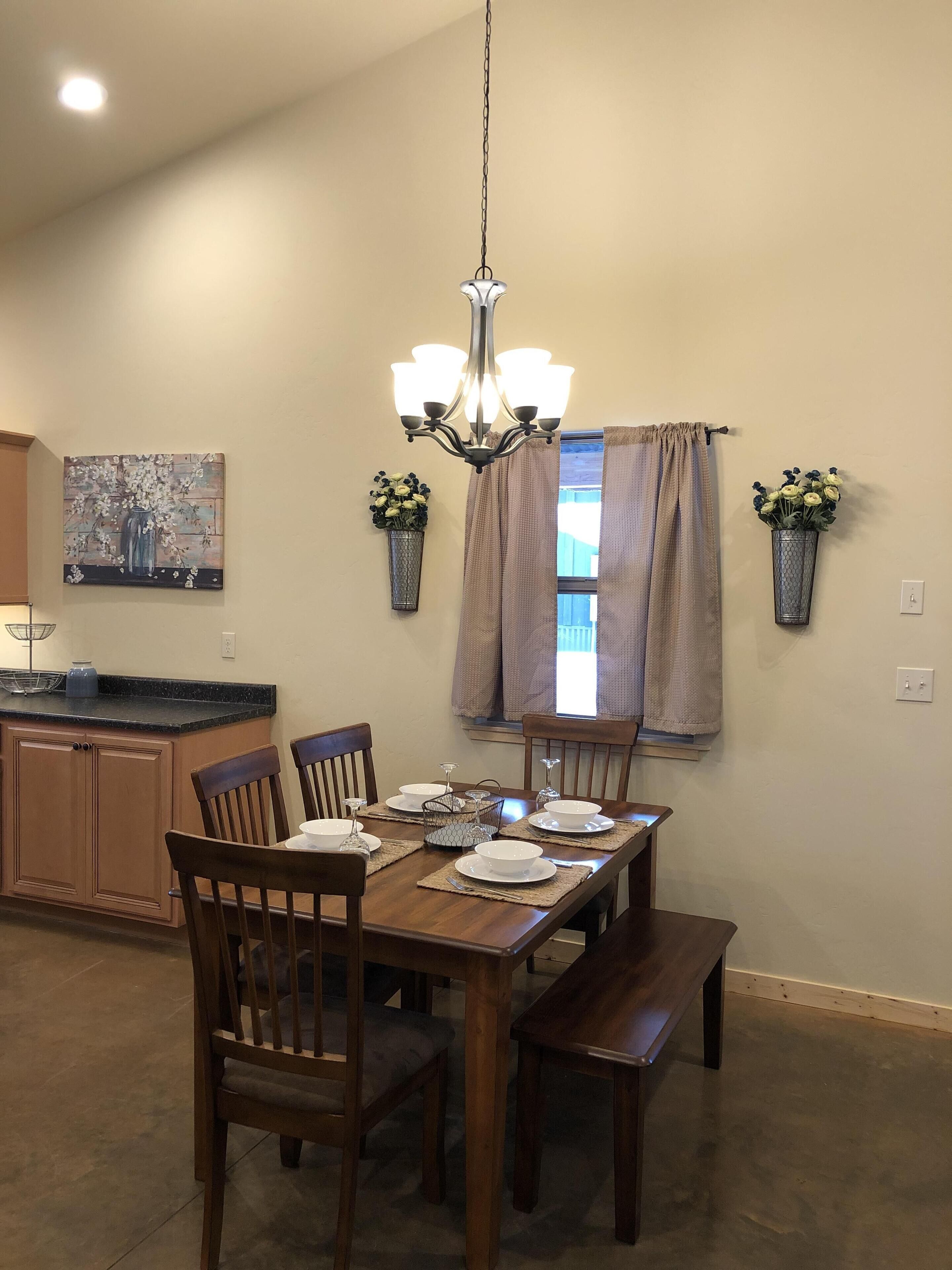 Un comedor con una mesa de madera dispuesta para cuatro personas, rodeada de sillas. Un candelabro cuelga del techo y hay dos plantas en macetas sobre las paredes.