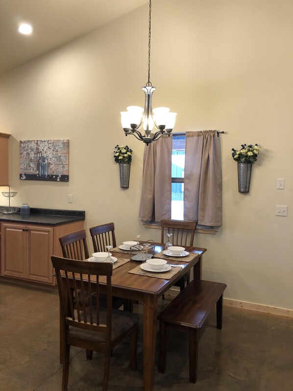 A dining area with a wooden table set for four, surrounded by chairs. A chandelier hangs from the ceiling, and there are two potted plants on the walls.