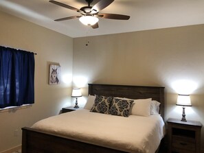 A bedroom with a bed, two bedside tables with lamps, a ceiling fan, a window with curtains, and a wall-mounted picture.