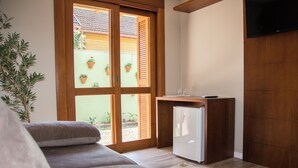 A room with a sofa, a small white refrigerator, a television, and a sliding glass door leading to an outdoor area with potted plants. - Pousada Caminho dos Plátanos (Gramado)