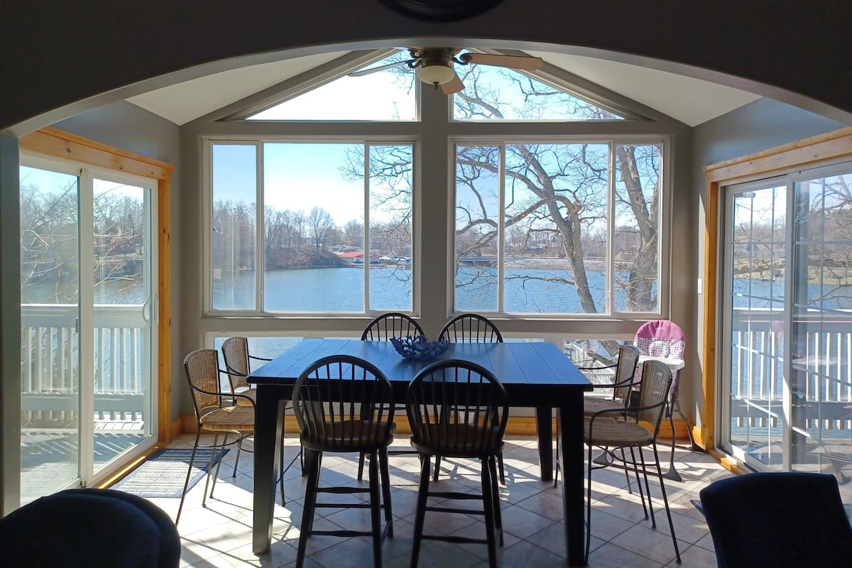 Ein Wintergarten mit großem Fenster mit Blick auf einen See, einem Esstisch mit Stühlen und einem Deckenventilator.