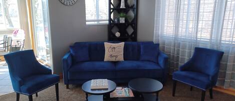 A living room with a blue sofa, two blue armchairs, a coffee table, and a bookshelf with decorative items.