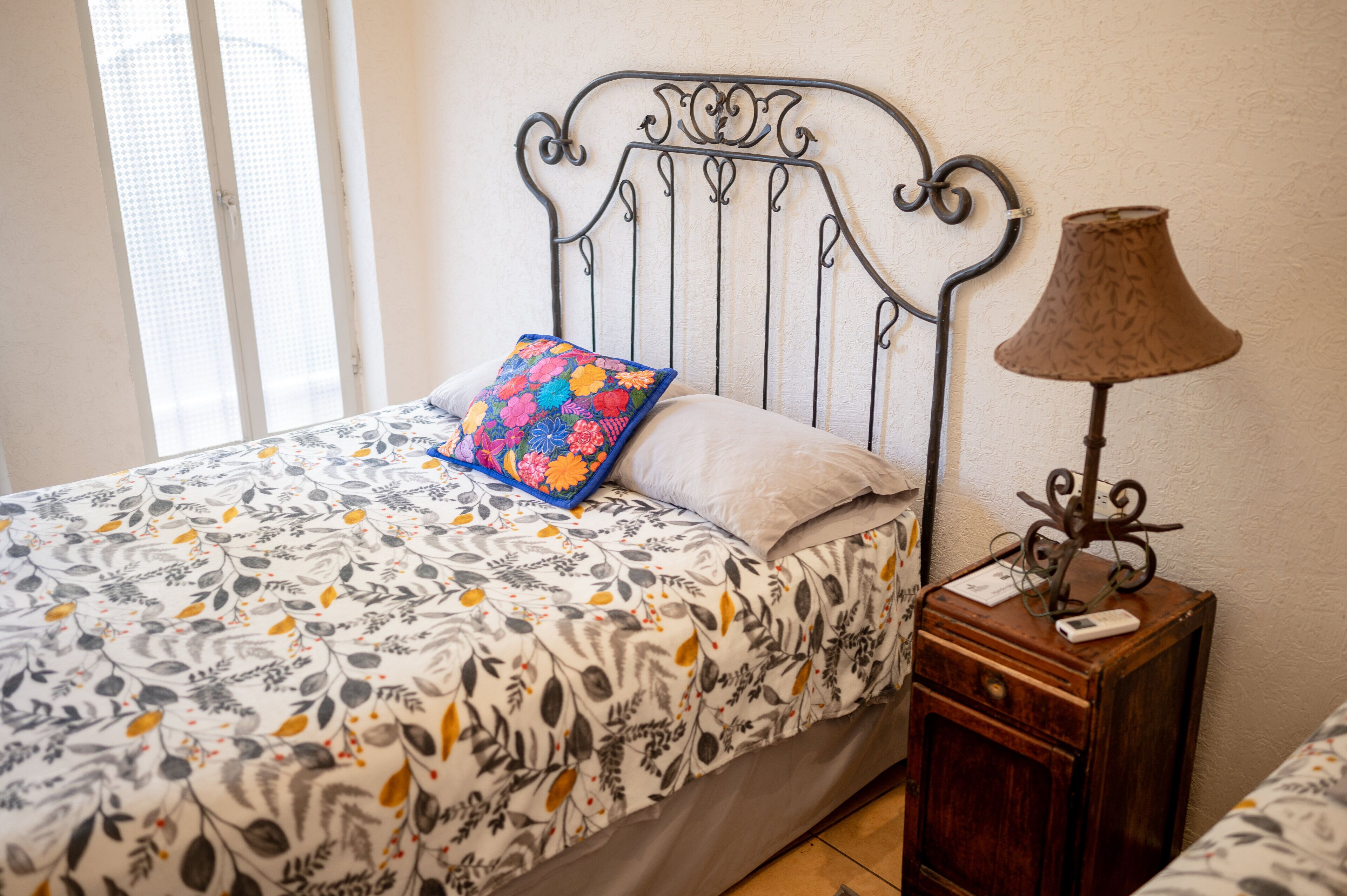 A bed with a patterned bedspread, a decorative pillow, a bedside lamp, and a small wooden nightstand.