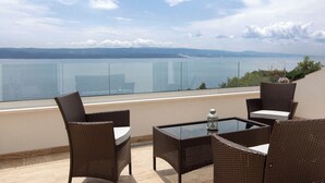 Ein Balkon mit Korbmöbeln und einem Glastisch, mit Blick auf ein Gewässer und ferne Berge.