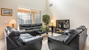 A modern living room with a black leather sofa, a coffee table, a TV, and a large window with blinds. - 7BR Pool Home Emerald Island by SHV-2793 (Kissimmee)