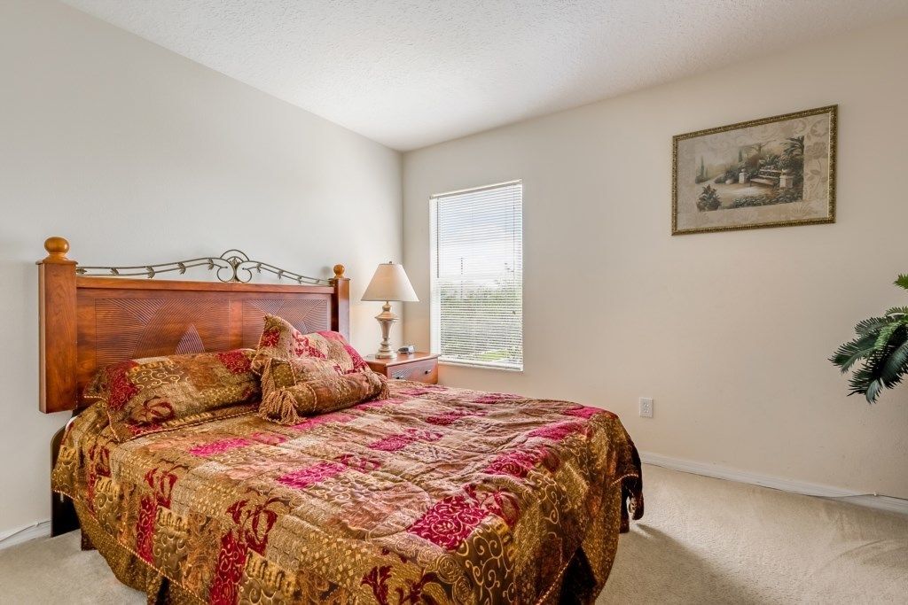 A bedroom with a large bed, a nightstand, a lamp, a window with blinds, and a framed picture on the wall.