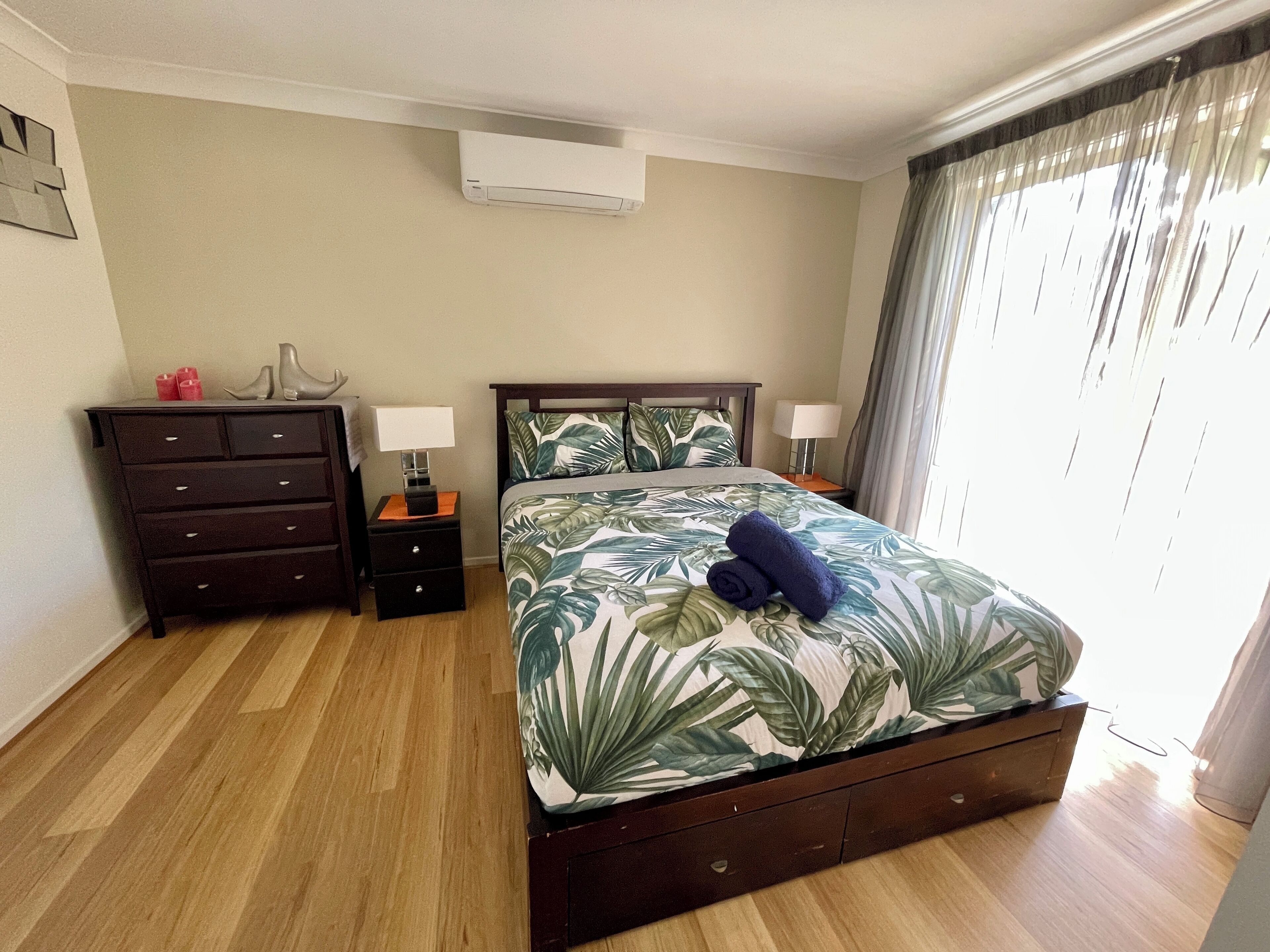 A hotel room with a bed, a dresser, two bedside tables with lamps, a window with curtains, and an air conditioning unit on the wall.