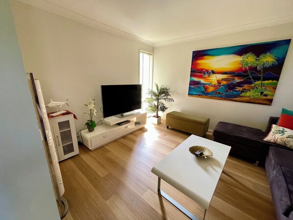 Un salon avec un téléviseur à écran plat, une petite table, un canapé, une plante en pot et un tableau accroché au mur.