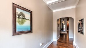 Un pasillo con suelo de madera, un cuadro de paisaje enmarcado en la pared izquierda y un candelabro al fondo.