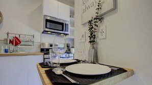 Una cocina con armarios blancos, un microondas y una estufa. Hay una mesa de comedor dispuesta con platos, vasos y cubiertos. Hay una planta en la pared y se ve un letrero con texto.