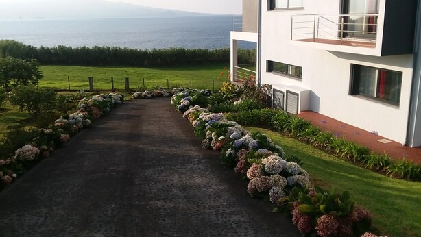 A modern two-story building with a paved driveway leading to a gated entrance, surrounded by lush greenery and a view of the sea. - Azores Beach Home for adults/children, 3 bed.2 bath, First Fl., 3534/AL (Horta, Azores)
