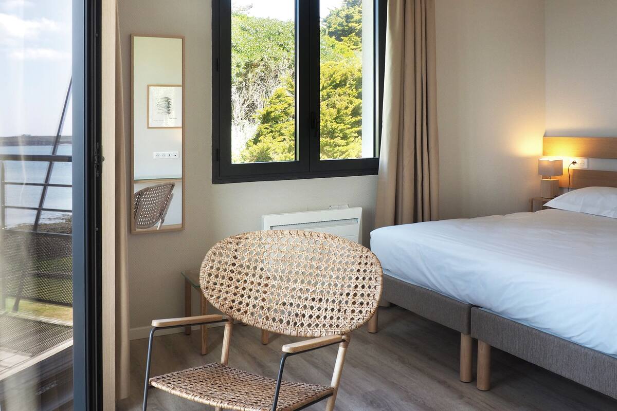 A hotel room with a bed, a rocking chair, a window with a view of trees, a ceiling light, and a wall-mounted air conditioner.